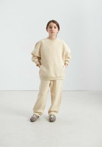 Sweat-shirt en polaire beige avec épaules tombantes et poche avant, assorti à un pantalon de survêtement beige et des chaussures à imprimé animalier.