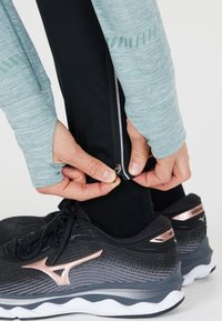 Urheilukengät mustana, joissa on hopeisia ja ruusukultaisia yksityiskohtia. Henkilö säätää vetoketjua mustissa urheiluleggingeissä. Huomiota herättävä kangas näkyy.