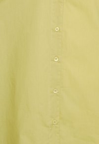 Camicia di cotone giallo chiaro con una trama liscia, dotata di una fila di sei bottoni rotondi sul davanti per la chiusura.