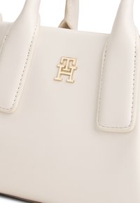 Τσάντα χειρός σε κρεμ απόχρωση με συνθετικό υλικό και λεία υφή, διαθέτει διπλές κοντές λαβές και μεταλλικό λογότυπο στο μπροστινό μέρος.