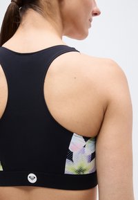 Femme portant un soutien-gorge de sport noir dos nageur avec des panneaux latéraux floraux, vue de dos sur un fond clair uni.