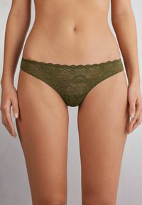 String en dentelle vert olive avec motif floral, taille scallopée et texture transparente. Conçu avec une couverture minimale et des détails délicats.