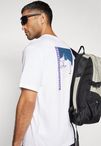 Camiseta blanca de manga corta con un gráfico de montaña azul y el texto "NUNCA DEJES DE EXPLORAR" en la parte posterior; mochila negra y gris.