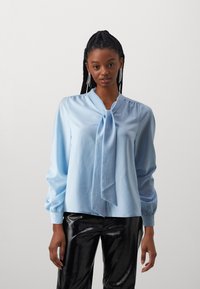 Lyseblå bluse laget av glatt stoff, med en knyttet hals og lange puffermer. Kombinert med skinnende svarte, tettsittende bukser.