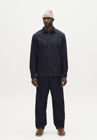 Marineblauwe denim shirt en bijpassende broek, beide met een relaxte pasvorm. Bruine leren schoenen. Ongerolde beige gebreide beanie op het hoofd.