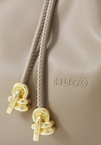 Material em pele bege com o logótipo HUGO em relevo e dois cordões de ajuste bege com terminais metálicos em forma de coração dourado brilhante, gravados com HUGO.