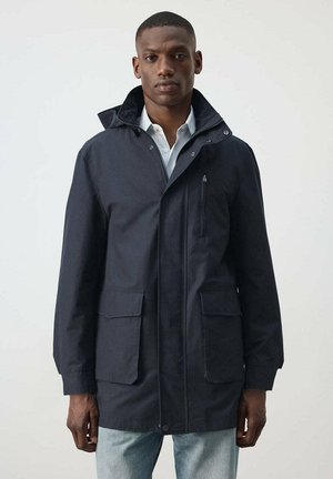 Hombre con una chaqueta con capucha de color azul marino oscuro sobre una camisa blanca y vaqueros azul claro, de pie frente a un fondo liso y claro.