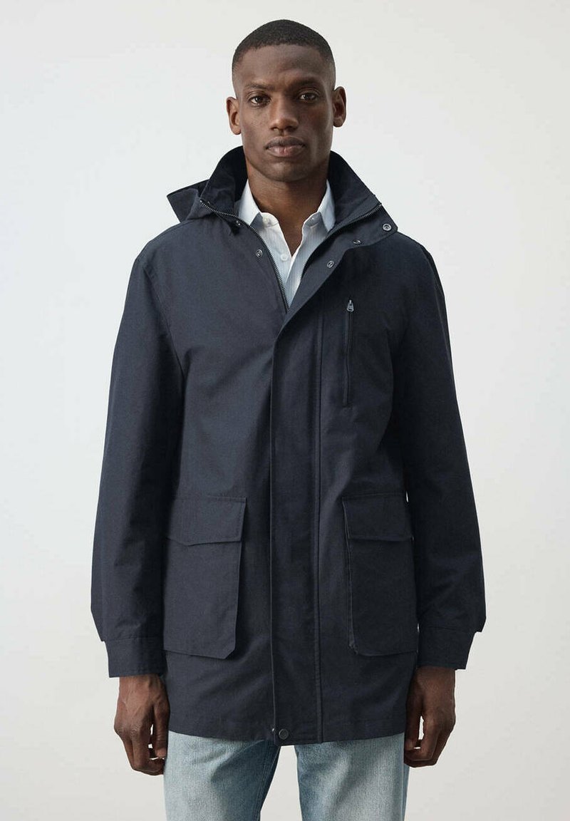 Homme portant une veste à capuche bleu marine foncé sur une chemise blanche et un jean bleu clair, debout devant un fond clair uni.