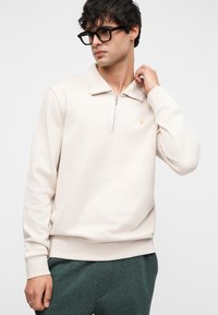 Μπεζ πουλόβερ με φερμουάρ, φαρδύ γιακά, ριμπ μανσέτες και τελείωμα. Διαθέτει μικρό κίτρινο λογότυπο στο στήθος. Απαλή, λεία υφή.