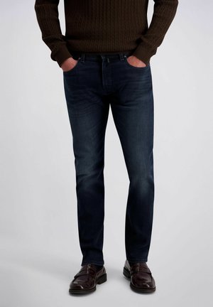 Mann trägt dunkelblaue Jeans, braunen Pullover und dunkelbraune Lederschuhe, steht mit den Händen in den Taschen vor einfachem Hintergrund.