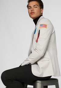 Light gray blazer with badge patches and red zipper detail, paired with dark gray fitted pants, set against a plain background.