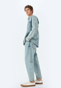 Giacca di jeans azzurro chiaro e jeans, vestibilità rilassata, con taschino sul petto, leggermente vissuto, e sneakers crema con suola color beige.