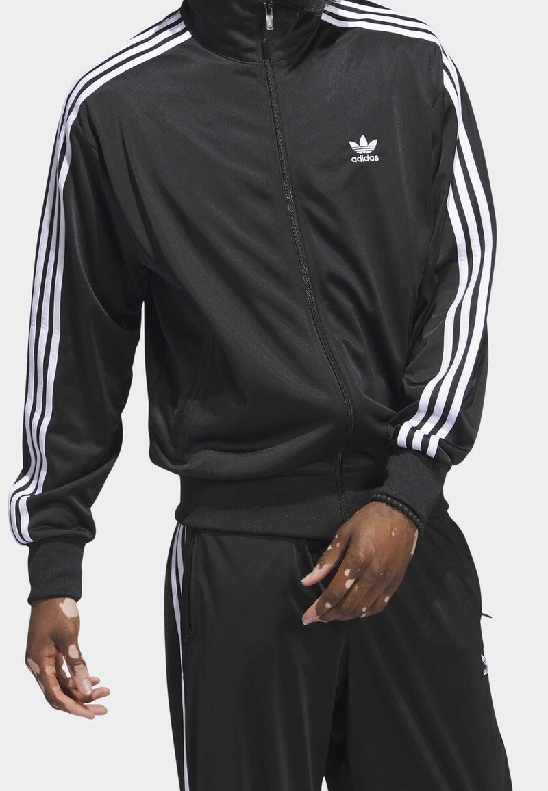 Černé polyesterové teplákové soupravy s bílými třemi pruhy na rukávech a nohavicích. Obsahuje zapínání na zip vpředu a žebrované manžety. Logo Adidas na hrudi.