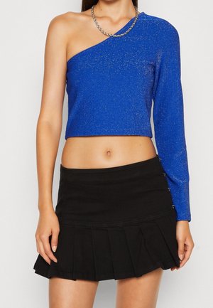 Kvinde iført en blå crop top med ét ærme og langærmet samt en sølvkædehalskæde, kombineret med en sort plisseret mininederdel.