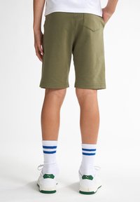 Shorts en coton vert olive avec une coupe décontractée, des coutures visibles et des poches latérales. Associés à des chaussettes blanches à rayures bleues et des baskets blanches.