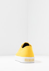 Żółty trampek z płótna z białą gumową podeszwą, o prostym designie, przeszyciach dekoracyjnych i logo na pięcie.