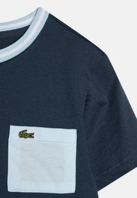 T-shirt en coton bleu marine avec une poche bleu clair. Présente un logo de crocodile vert sur la poche. Col côtelé en bleu clair et blanc.