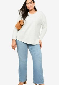 Ljusgrå långärmad V-ringad topp i kombination med ljusblå raka jeans. Modellen håller en brun mockaväska med subtila texturer.