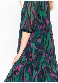 Plovoucí šaty s abstraktním vzorem v zelené a černé barvě s fialovými akcenty. Mají vrstevnatou sukni a nafouknuté rukávy. Lehce přenosný materiál.