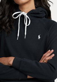 Černá mikina s kapucí vyrobená z texturovaného materiálu, která má bílé šňůrky a malé bílé logo polo na prsou. Standardní střih s žebrovanými manžetami.