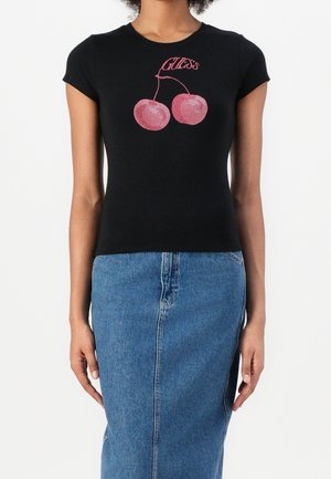 T-shirt noir ajusté à manches courtes avec des cerises en strass rouges et le texte "Guess" au-dessus. Associé à une jupe en jean bleue.