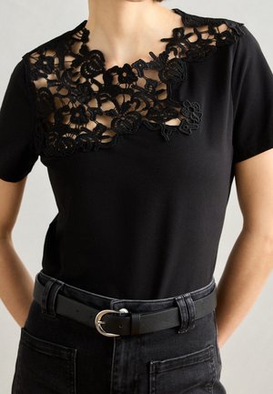 Haut noir à manches courtes avec détail en dentelle florale sur le haut de la poitrine et de l'épaule, associé à une ceinture noire et un jean foncé.