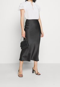 Kvinna iförd en vit volangblus med kort ärm, svart satinmidikjol, svarta klacksandaler och håller en svart handväska med ett metallhandtag.