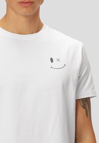 T-shirt en coton blanc avec un col rond, présentant un graphique de smiley gris sur la poitrine gauche, texture lisse et manches courtes.