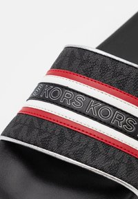 Sandale à glissière noire avec une sangle texturée présentant des rayures rouges, blanches et noires et la marque "KORS" en lettres blanches.