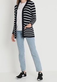 Granatowo-biały kardigan w paski, z długimi rękawami, z otwartym przodem, noszony na jasnym t-shircie. Połączony z jasnoniebieskimi dżinsami i czarnymi trampkami.