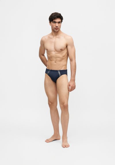 Μυώδης άνδρας στέκεται ξυπόλυτος με το πρόσωπο προς τα εμπρός, φοράει σκούρα μπλε εσώρουχα με ελαφριά κατακόρυφη ραφή σε απλό λευκό φόντο.