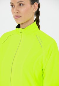 Neon yellow athletic jacket with a high collar, front zipper, and reflective accents. Features a textured fabric and fitted design.
