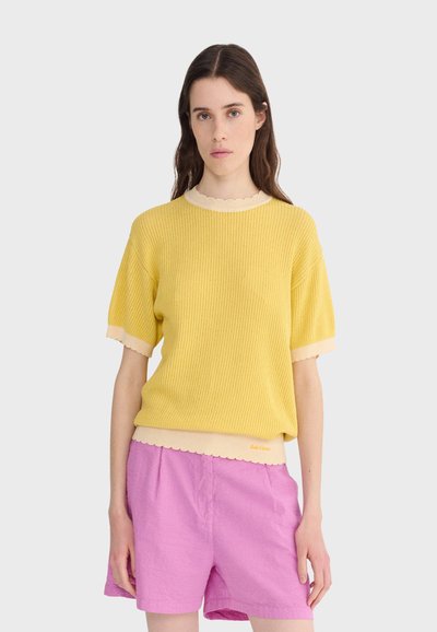 Femme aux longs cheveux bruns portant un pull jaune à manches courtes avec bordure beige et un short taille haute rose, debout devant un fond uni.