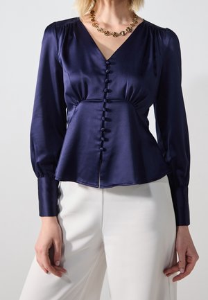 Vrouw die een marineblauwe satijnen blouse draagt met knopen bedekt met stof en witte broek, geaccentueerd met een gouden ketting.