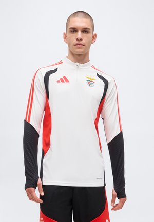 Giovane uomo che indossa una maglia sportiva bianca con cerniera a quarto, con dettagli rossi e neri e lo stemma del club di calcio Benfica, in piedi su sfondo semplice.