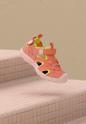 Une sandale pour enfant Viking corail et jaune flotte au-dessus de marches carrelées, devant un mur beige.