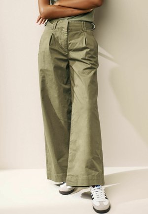 Persona con pantalones anchos de color verde oliva y zapatillas blancas con rayas negras, de pie con los brazos cruzados frente a una pared beige.