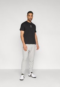 Schwarzes T-Shirt mit kurzen Ärmeln und weißem Logo, kombiniert mit hellgrauen Jogginghosen und weißen Sneakern mit schwarzen und goldenen Akzenten.