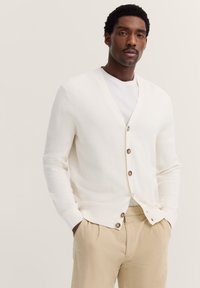 Weiße Strickjacke mit V-Ausschnitt, braunen Knöpfen, gerippten Bündchen und lockerer Passform, getragen über einem weißen T-Shirt und beigen Hosen.