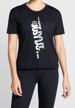 Frau trägt ein schwarzes kurzärmeliges Sportshirt mit weißem vertikalem "Just Do It." Text und schwarze Leggings vor grauem Hintergrund.
