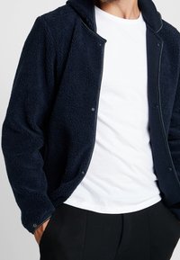 Marineblå fleecejakke med en struktureret overflade, trykknapper og en høj krave, lagd ovenpå en hvid t-shirt og sorte bukser.
