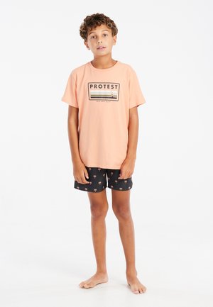 Lichtoranje t-shirt met het "PROTEST" logo en strepen, gecombineerd met zwarte shorts met kleine palmboompatronen. Jong persoon op blote voeten.