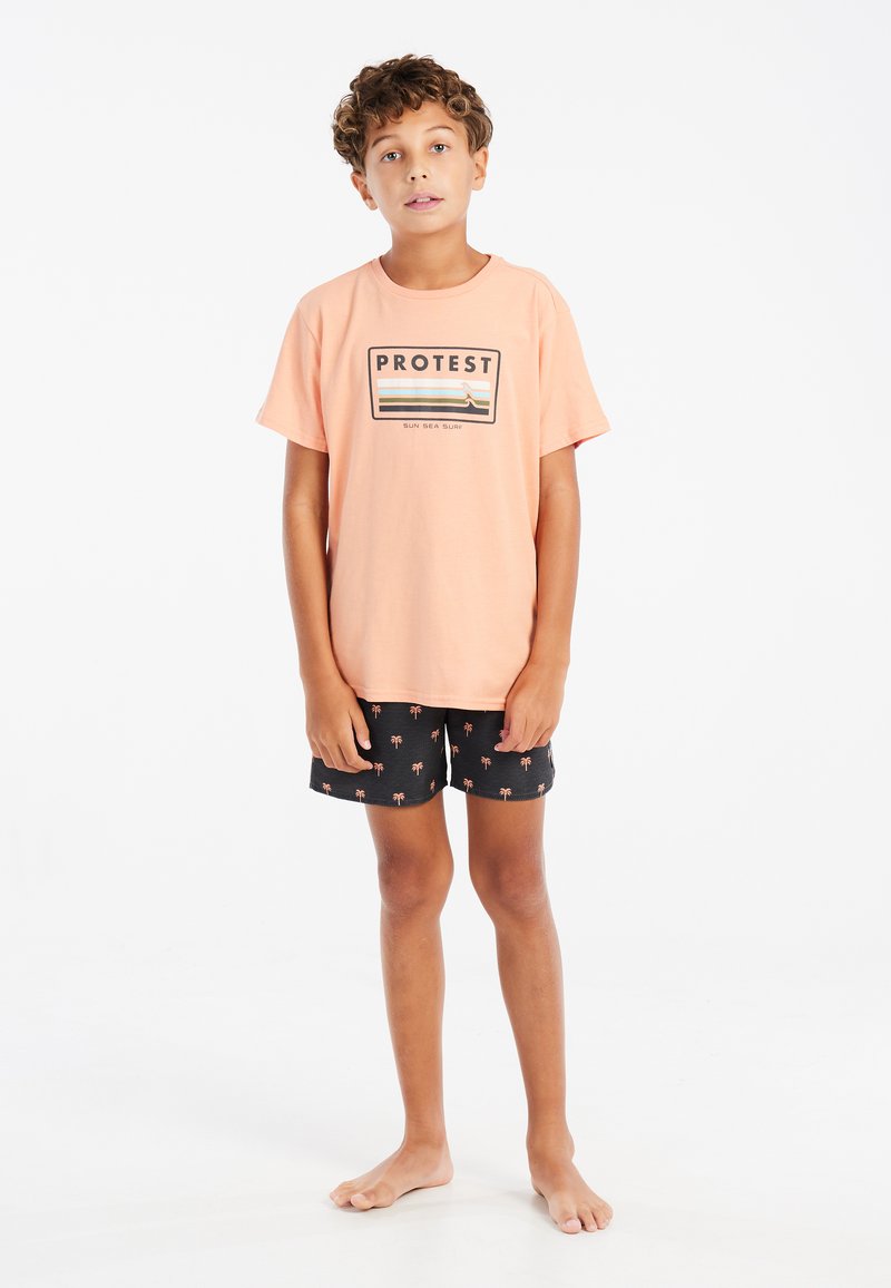 Lichtoranje t-shirt met het "PROTEST" logo en strepen, gecombineerd met zwarte shorts met kleine palmboompatronen. Jong persoon op blote voeten.