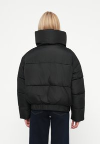 Geacă puffer neagră cu guler înalt, textură matlasată, tiv elastic și croială supradimensionată, purtată peste blugi de culoare bleu închis.