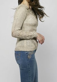 Femme portant un pull en maille beige avec des détails en dentelle sur les manches et un jean bleu avec des motifs floraux brodés sur la poche et la cuisse.