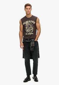 Camiseta gráfica sin mangas de color marrón con la impresión "Superdry Los Ángeles", jeans negros, camisa de cuadros atada a la cintura y mocasines negros.