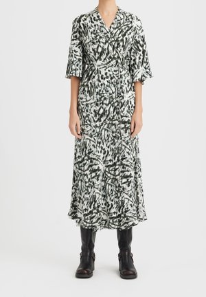 Robe longue de style enveloppant avec un motif abstrait vert et blanc. Elle présente un décolleté en V, des manches courtes et une taille cintrée. Portée avec des bottes noires.