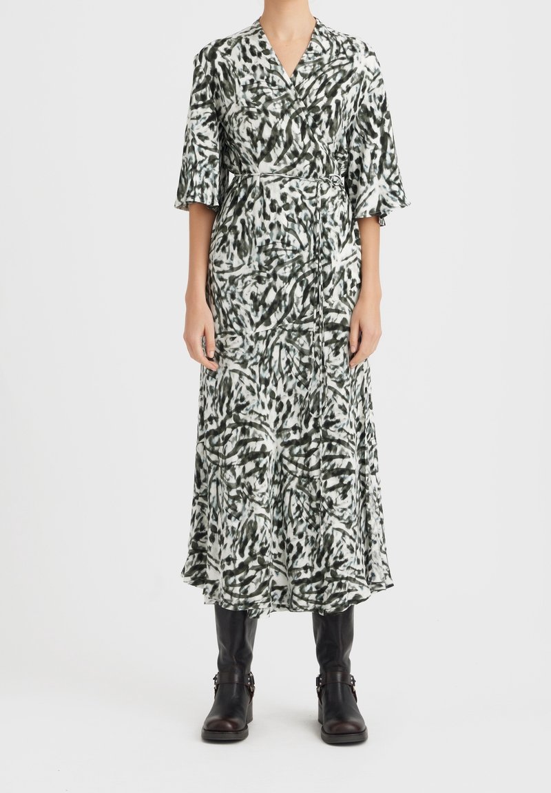 Robe longue de style enveloppant avec un motif abstrait vert et blanc. Elle présente un décolleté en V, des manches courtes et une taille cintrée. Portée avec des bottes noires.