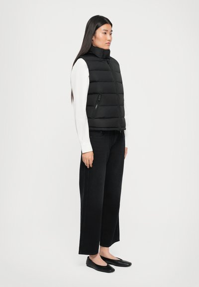Donna in piedi che indossa un gilet imbottito nero sopra una maglietta bianca a maniche lunghe, jeans neri a gamba larga e scarpe nere basse, su uno sfondo neutro.