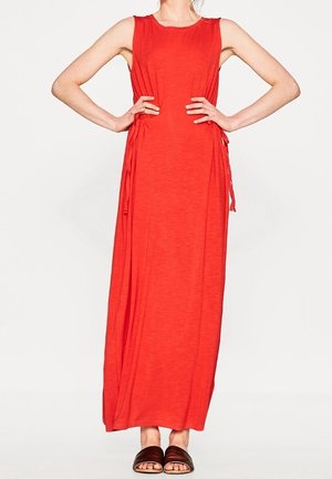 Bezrękawna, jasnoczerwona sukienka maxi z wiązaniami po bokach, noszona z brązowymi, otwartymi sandałami na płaskiej podeszwie, modelka stojąca z rękami na biodrach.